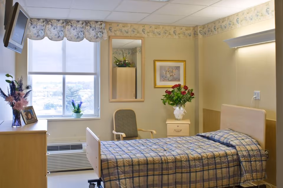 A small, neatly arranged bedroom in a senior living facility with a single bed covered in a plaid bedspread. There is a wooden nightstand with a vase of red and white flowers, a framed picture on the wall, a chair, a dresser with a photo frame and flower vase, and a window with floral curtains letting in natural light. A wall-mounted TV is visible in the upper left corner.