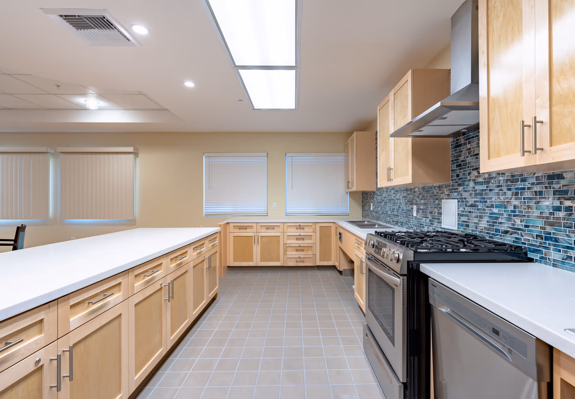 Bright modern communal kitchen with a long white island, light wood cabinets, stainless steel appliances and a blue mosaic tile backsplash.