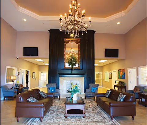 A spacious and elegant living room area with two brown leather sofas facing each other, a wooden coffee table in the center with a floral arrangement, and two armchairs with blue cushions. A large dark wood fireplace with a mirror and chandelier above it is the focal point of the room. The walls are painted beige, and there are two flat-screen TVs mounted on either side of the fireplace. Additional seating and decor are visible in the background.