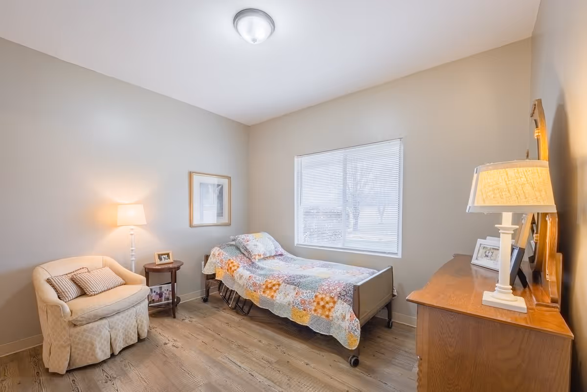 A small, cozy bedroom with a single bed covered in a colorful patchwork quilt. There is a beige armchair with two striped pillows, a round wooden side table with a framed photo, and a wooden dresser with a lamp and framed pictures. A window with blinds lets in natural light, and the walls are painted a soft beige color.