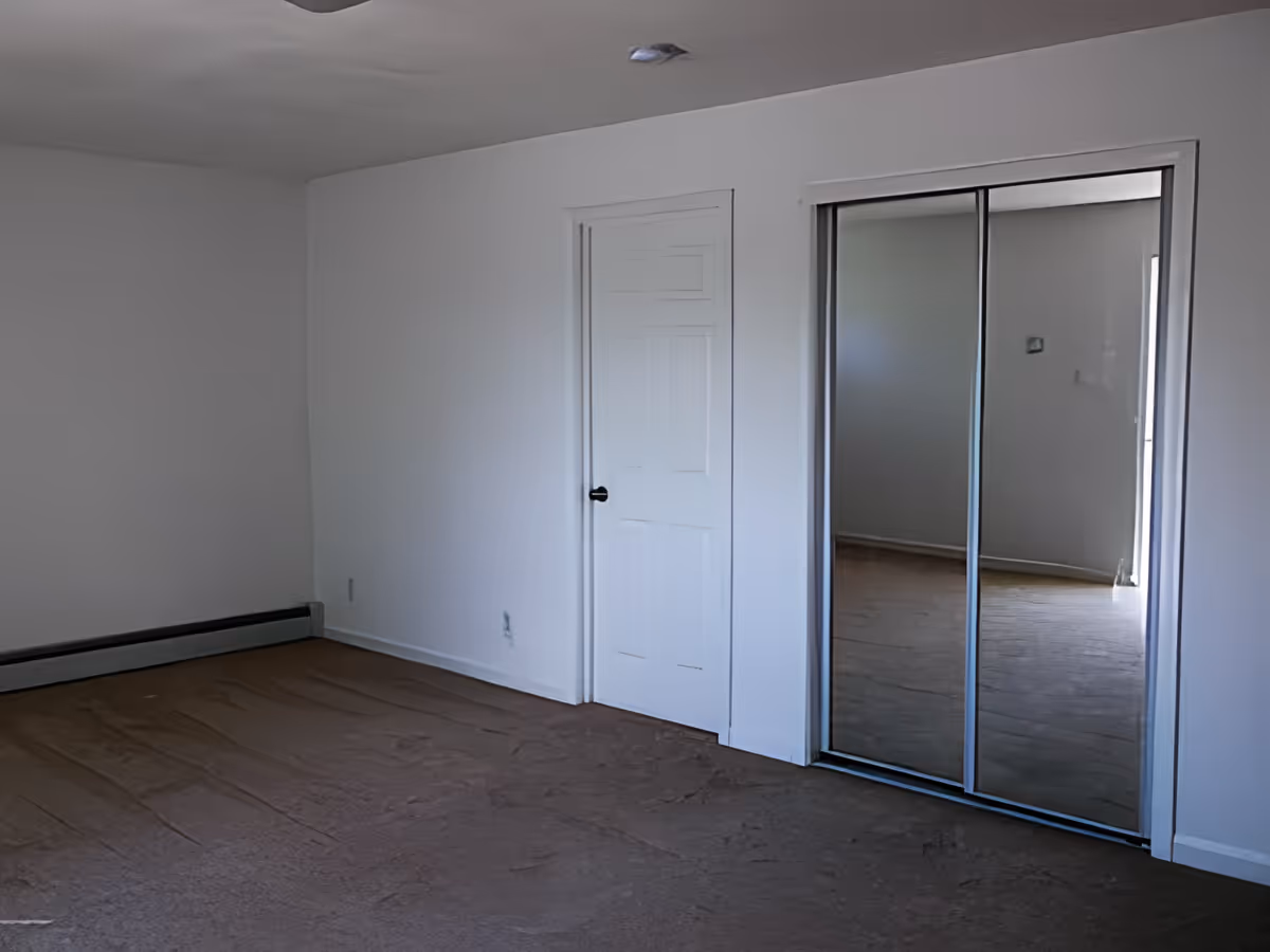 Empty room with white walls, a closed white door, and a closet with sliding mirrored doors reflecting part of the room. The floor is covered with brown carpet.