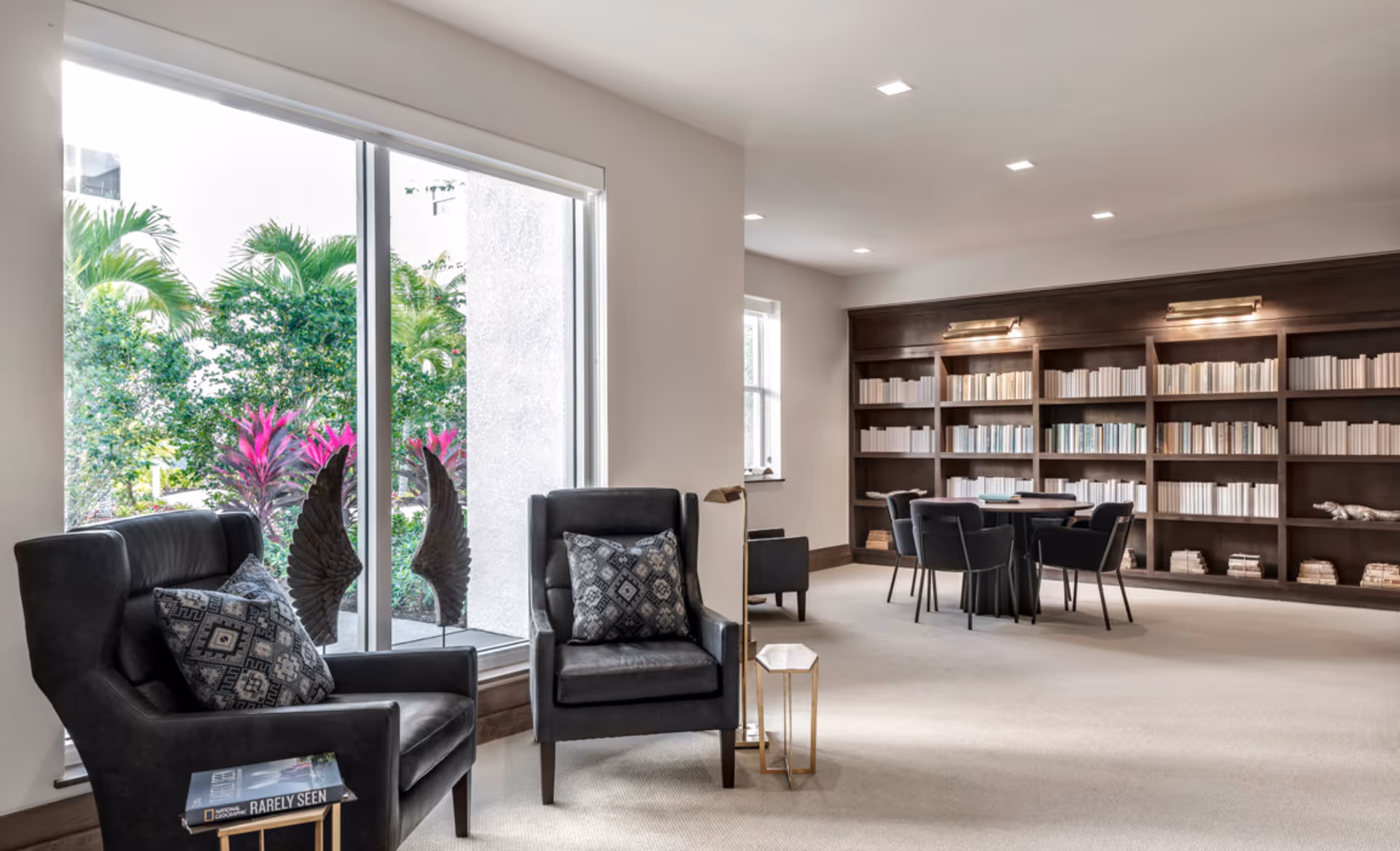 A bright and spacious reading room with two black armchairs featuring patterned cushions near a large window with a view of green plants outside. There is a small side table with a book on it next to one chair and a floor lamp between the chairs. In the background, there is a round table with four black chairs and a large dark wooden bookshelf filled with books.