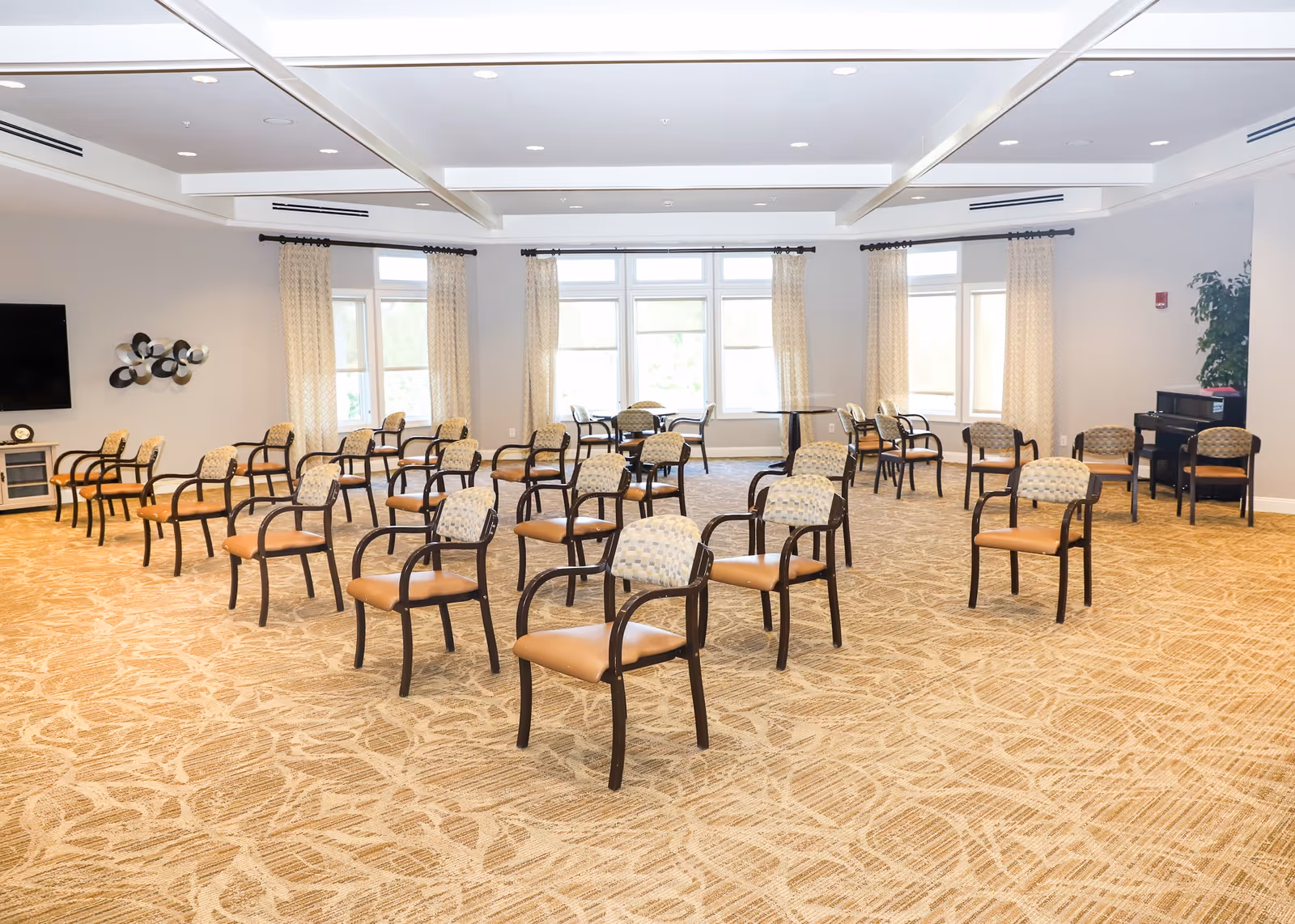 A spacious, well-lit room with multiple rows of chairs arranged facing forward, large windows with curtains allowing natural light, a wall-mounted TV on the left, and a piano with chairs on the right side of the room.