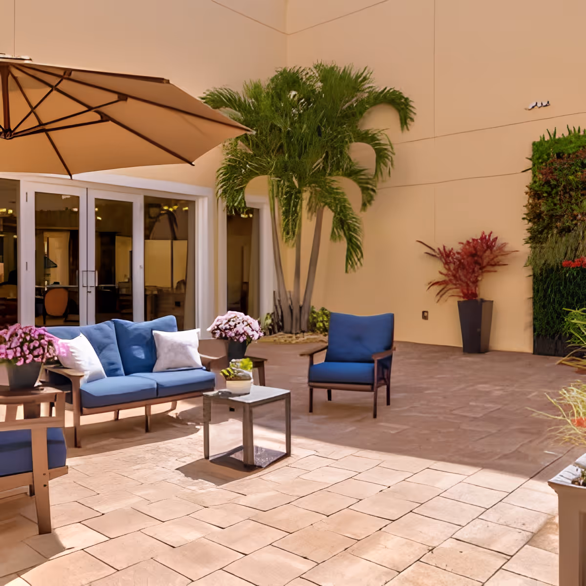 Outdoor patio area with beige tiled flooring, blue cushioned seating including a sofa and chairs, a small table, potted plants with pink flowers, a large beige umbrella, and tropical greenery against a beige building wall with glass doors.
