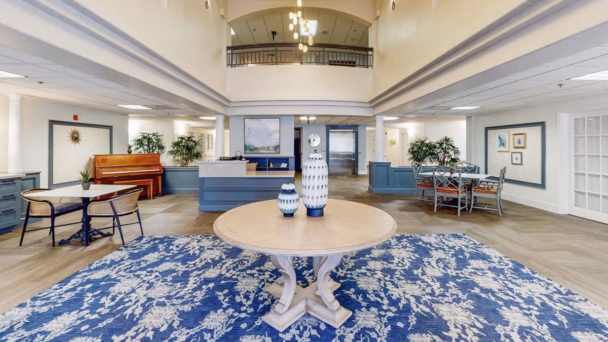 Spacious senior living lobby with a round wooden table on a blue patterned rug, seating areas, a piano, reception desk, and a double-height ceiling with balcony.