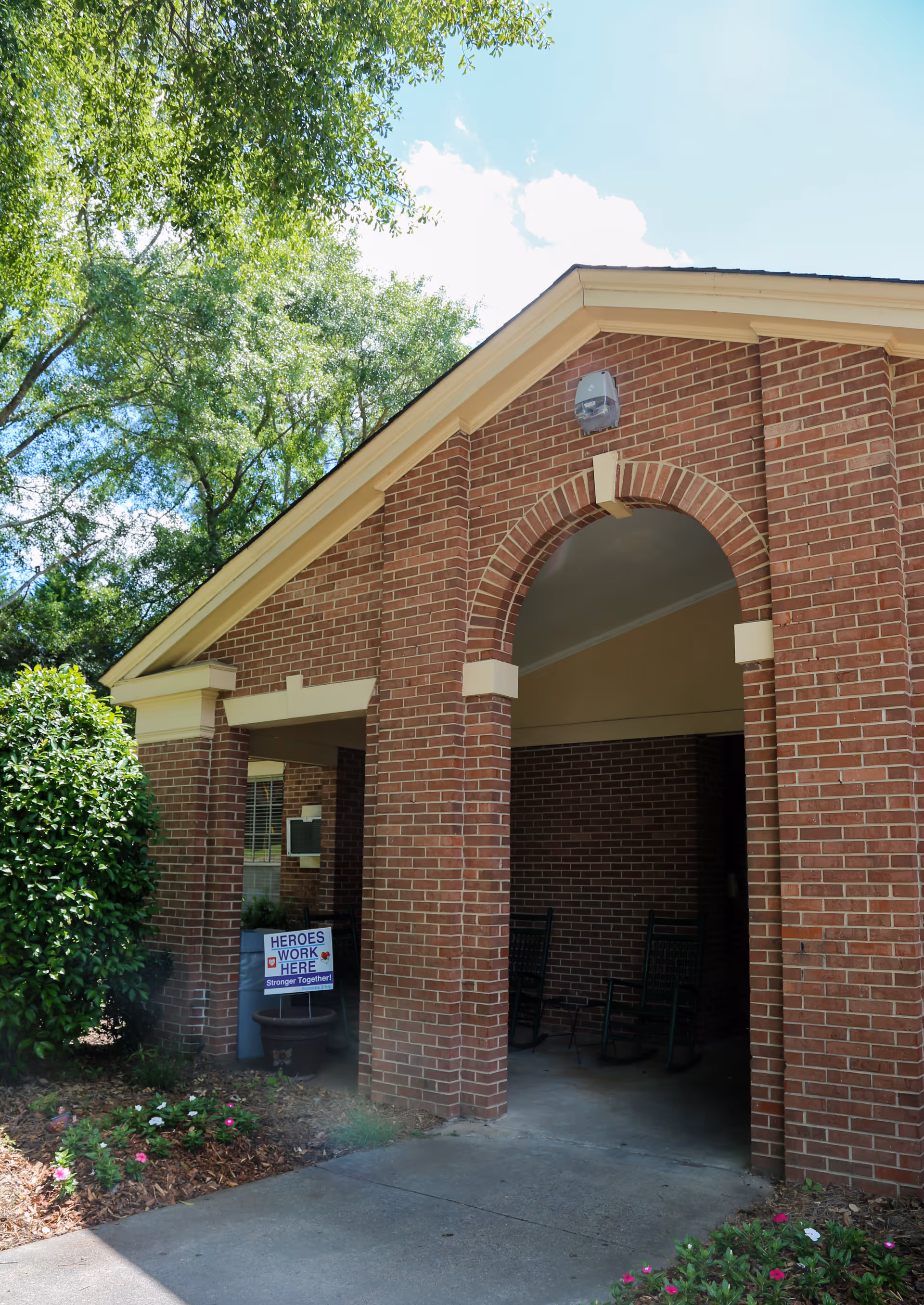 Entrance of a brick building with an arched doorway and a covered porch area containing rocking chairs. There are green bushes and flowers near the entrance, and a sign that reads 'HEROES WORK HERE Stronger Together!' is placed near the doorway. Trees and a blue sky with some clouds are visible in the background.
