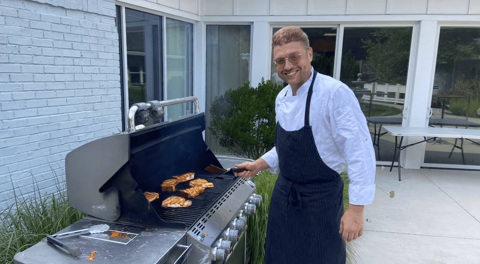 A man wearing a white chef's jacket and black apron is grilling several pieces of chicken on a large outdoor gas grill. He is smiling and holding a spatula. The setting appears to be an outdoor patio area with white brick walls, glass doors, and some greenery in the background.