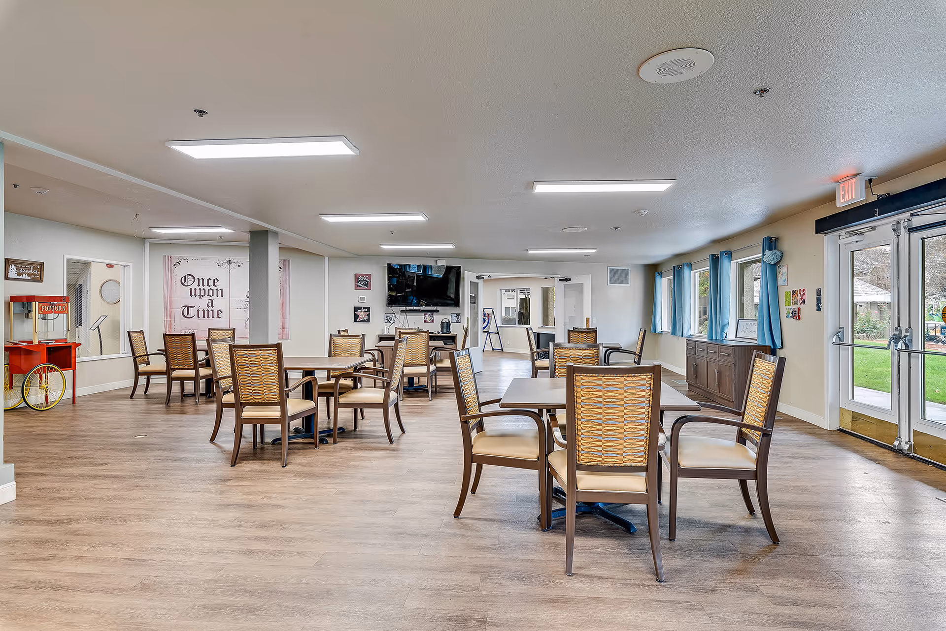 A spacious senior living dining room with multiple tables and chairs arranged neatly. The room has wood flooring, large windows with blue curtains, and a double glass door leading outside. On the left side, there is a red popcorn machine and a wall decoration with the text 'Once upon a Time'. A flat-screen TV is mounted on the far wall above a small cabinet.