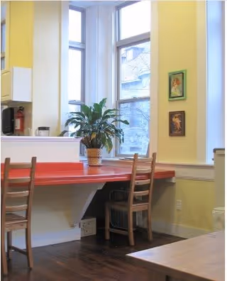 A bright interior corner with large windows letting in natural light. There is a red countertop with two wooden chairs placed in front of it. A potted green plant sits on the countertop. The walls are painted yellow and decorated with two small framed pictures. The floor is wooden.