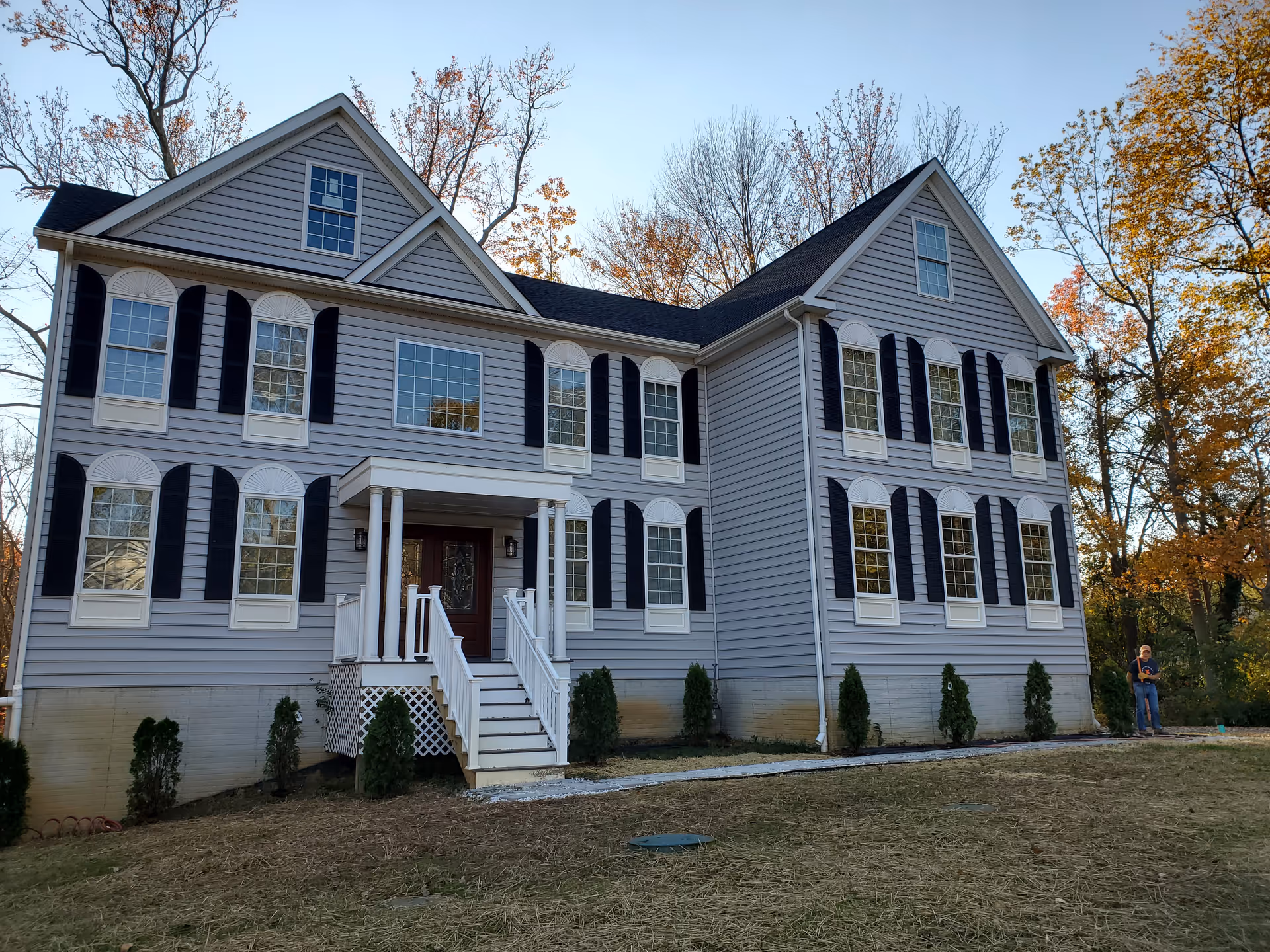 A large two-story gray house with white trim and black shutters, surrounded by trees with autumn foliage. There is a small porch with white railings and steps leading to a dark front door. A person stands near the right side of the house on the grass.