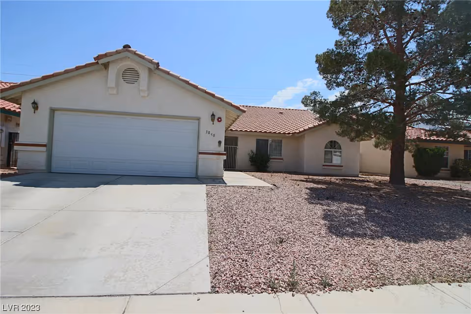 Single-story stucco house with a two-car garage, tile roof, rock landscaping and a large tree in the front yard.