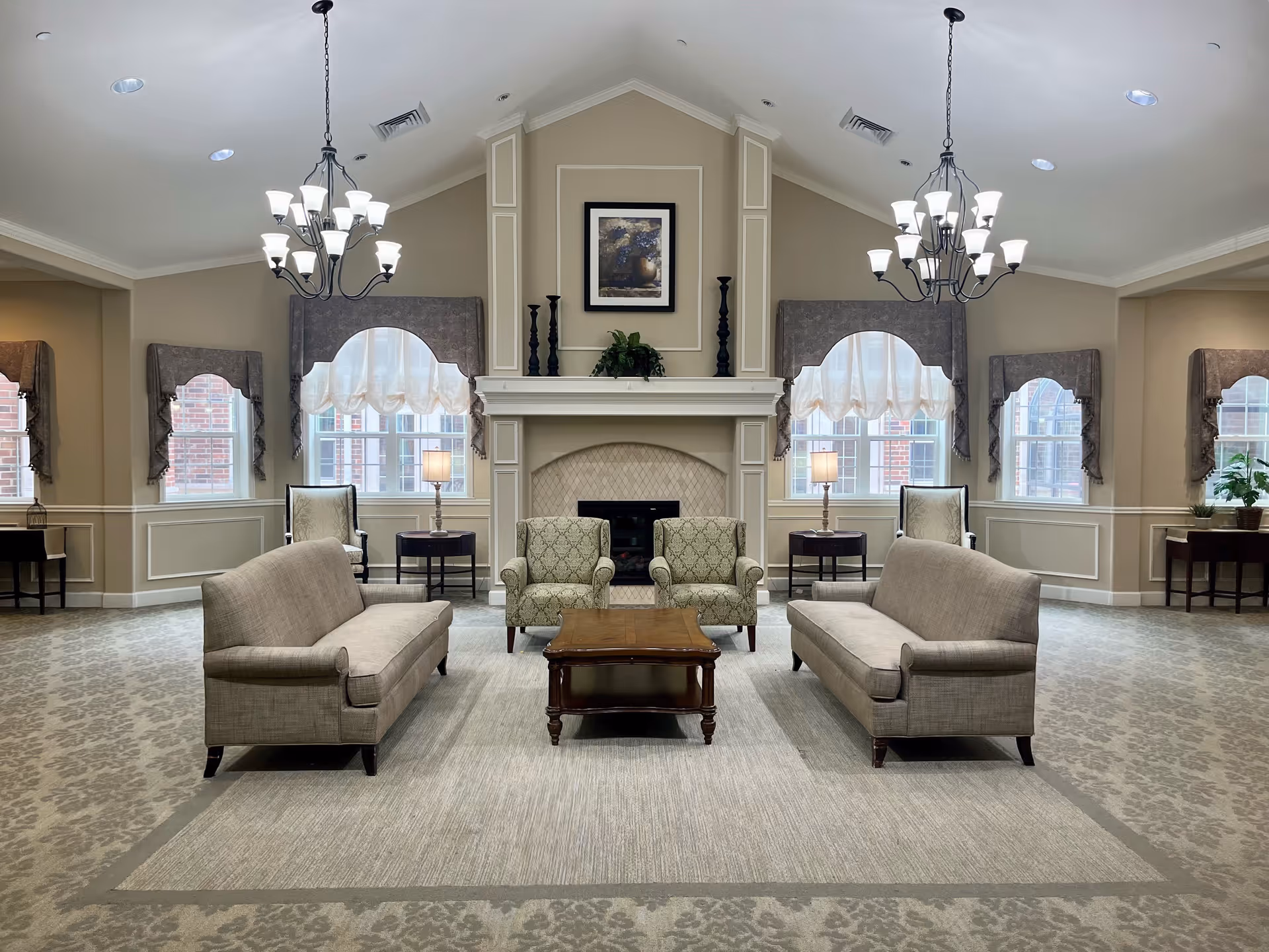 A spacious, well-lit living room area with two beige sofas facing each other, two patterned armchairs, and a wooden coffee table in the center. The room features a fireplace with decorative candlesticks and a framed picture above it. Large windows with sheer curtains and valances allow natural light to fill the space. Two chandeliers hang from the ceiling, and side tables with lamps are placed near the armchairs. The walls are painted beige with white trim, and the floor is covered with a patterned carpet and a large area rug.