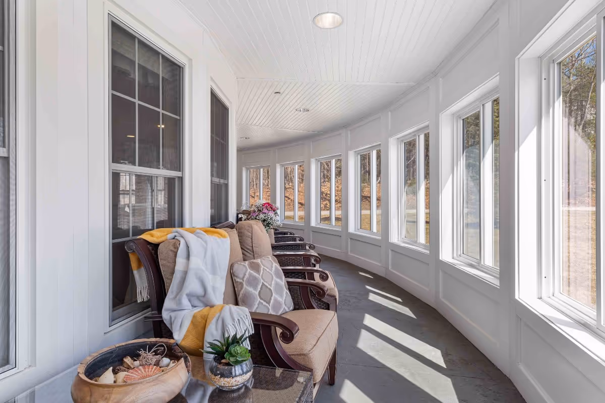 A bright, curved sunroom with large windows letting in natural light. The room features cushioned wooden armchairs with decorative pillows and a throw blanket. A small table holds a bowl with seashells and a small potted plant. Outside, trees and grass are visible through the windows.