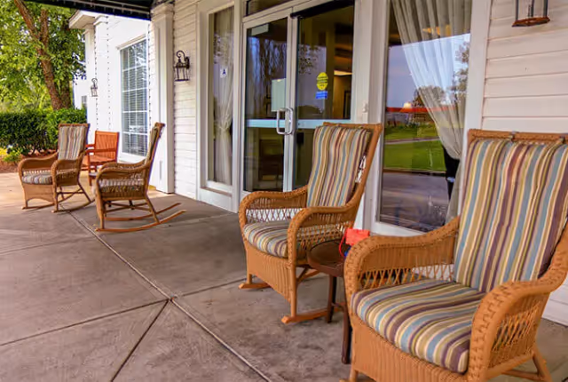 Outdoor patio area at Florence Place with several wicker rocking chairs featuring striped cushions arranged along the building's exterior wall near glass doors and windows, with greenery visible in the background.