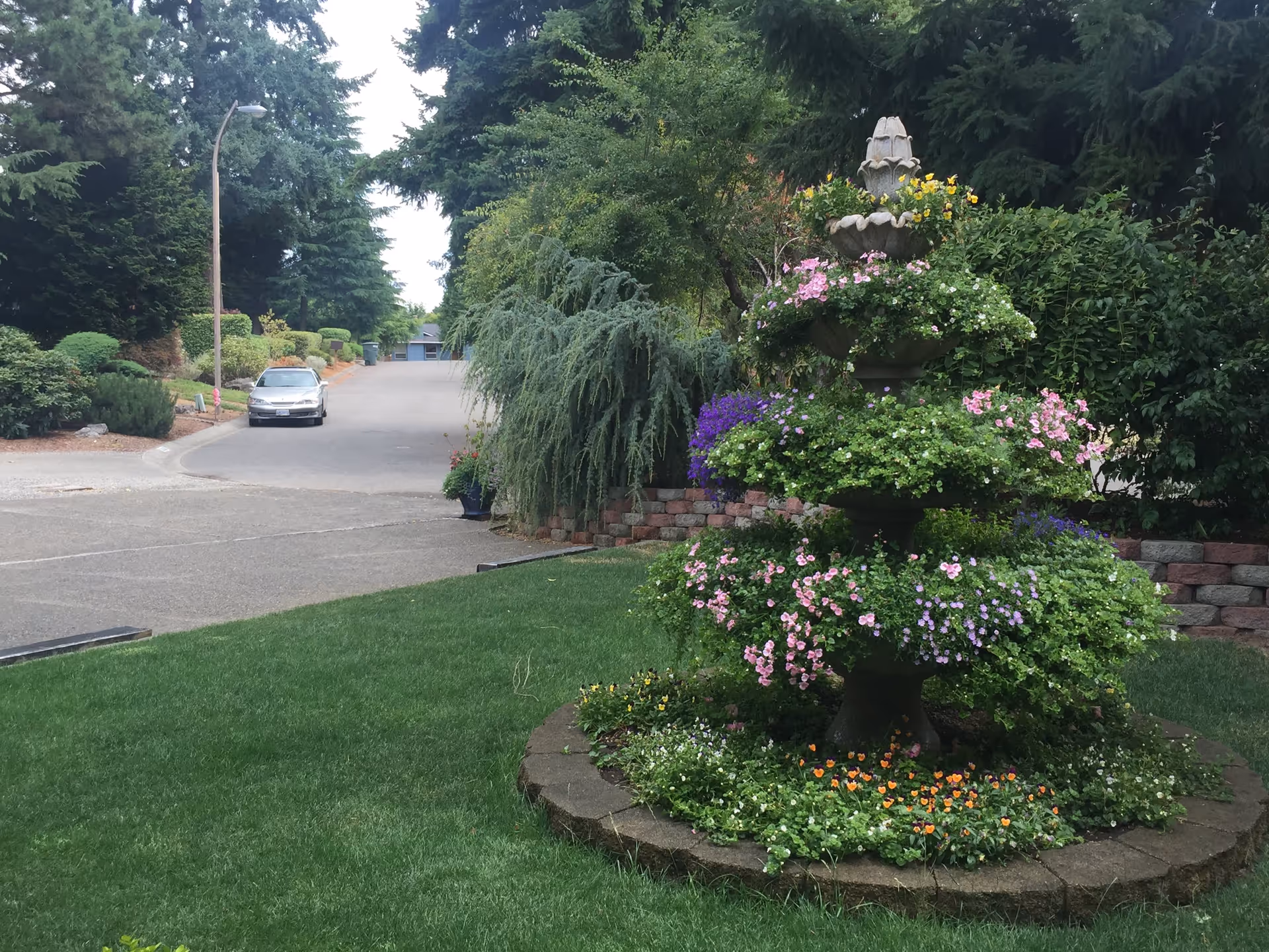 A landscaped outdoor area featuring a tiered stone fountain planter filled with various colorful flowers and greenery. The fountain is surrounded by a circular stone border and lush green grass. In the background, there is a paved road with a parked car, trees, and shrubs lining the street.