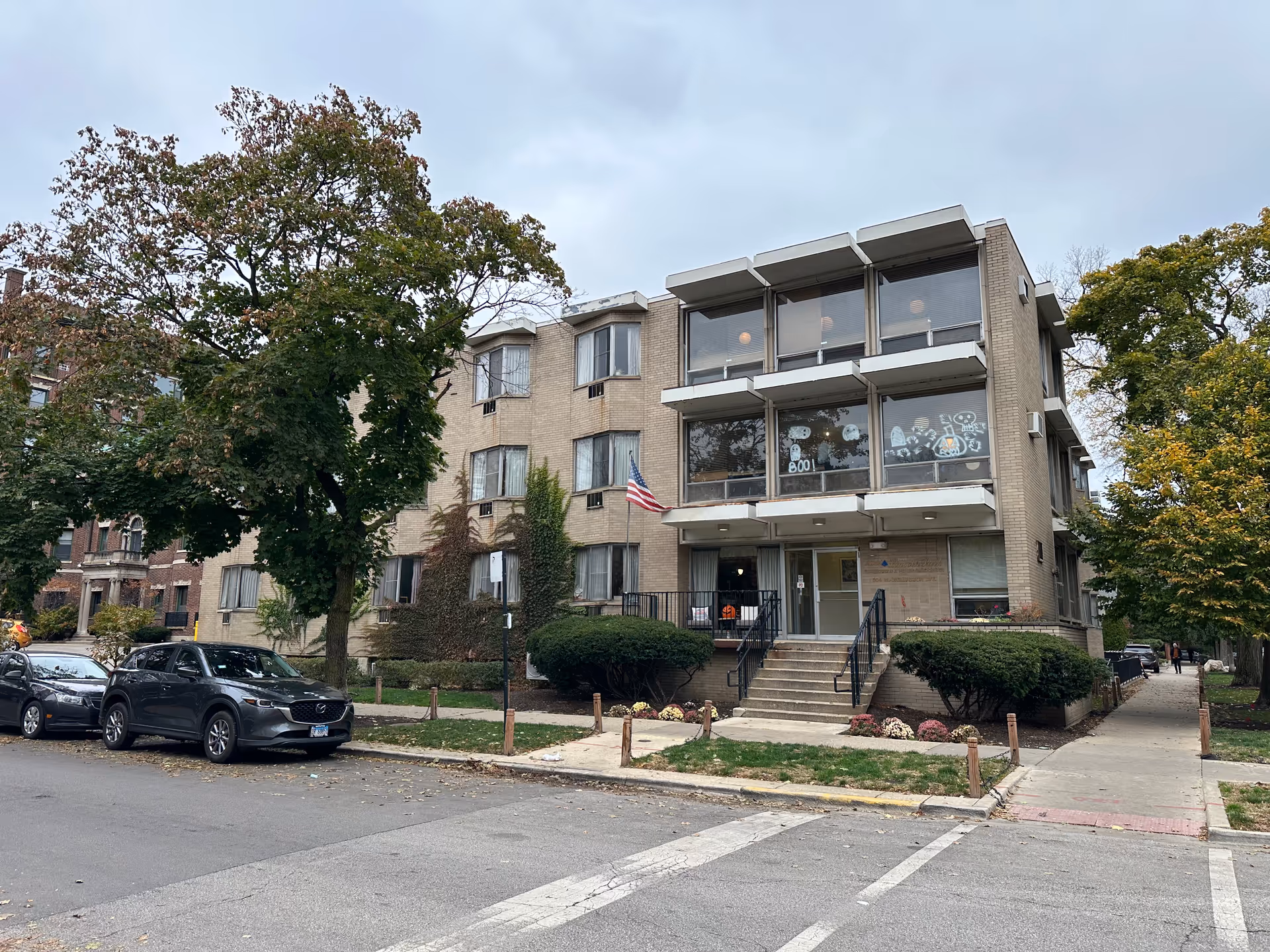 Exterior view of a three-story brick building with large windows decorated with Halloween-themed drawings. There are stairs leading up to the entrance, bushes, and trees around the building. Several cars are parked along the street in front of the building under a cloudy sky.