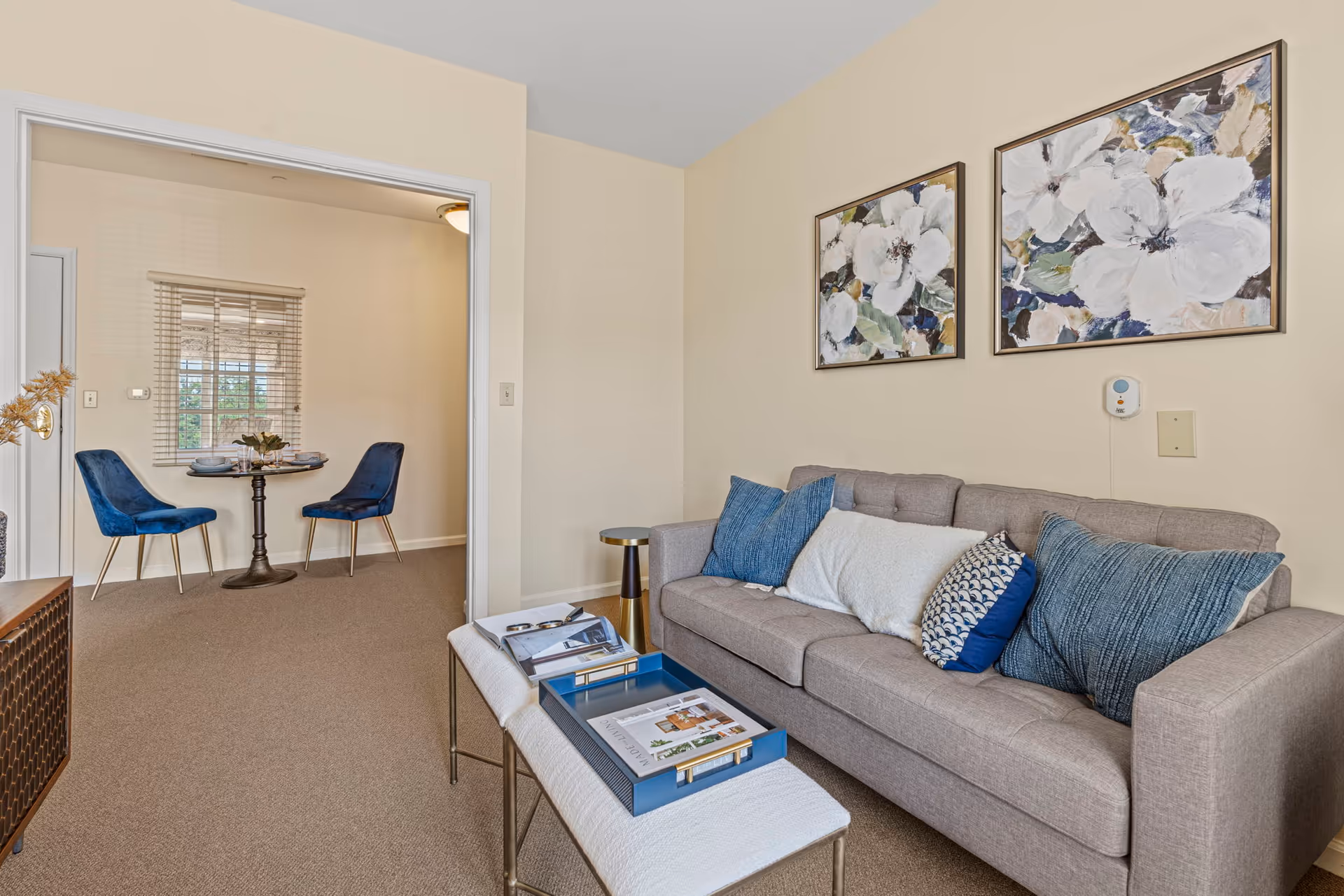A cozy living room with a gray sofa adorned with blue and white pillows, two floral paintings on the wall above the sofa, and a small bench with a blue tray and magazines. Through an open doorway, a small dining area is visible with a round table set for two and two blue velvet chairs near a window with blinds.