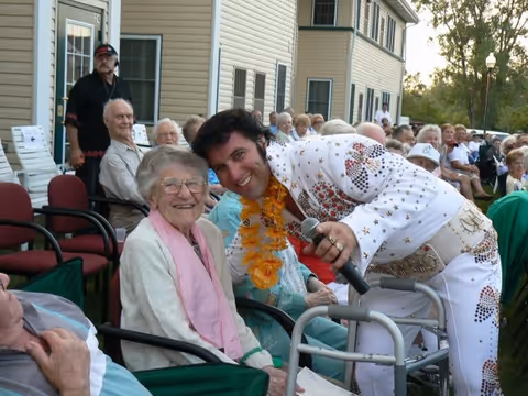 An elderly woman sitting outdoors in a chair smiling while a man dressed in an Elvis Presley-style white jumpsuit with colorful decorations and a yellow lei leans toward her holding a microphone. Other elderly people are seated in rows of chairs outside a building, watching the interaction.