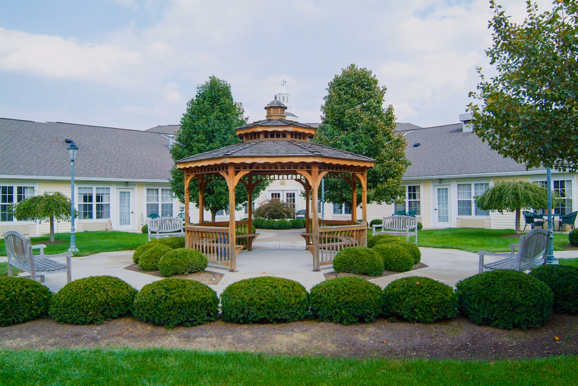 A wooden gazebo sits in a landscaped courtyard with benches, trimmed shrubs, and a single-story building in the background.