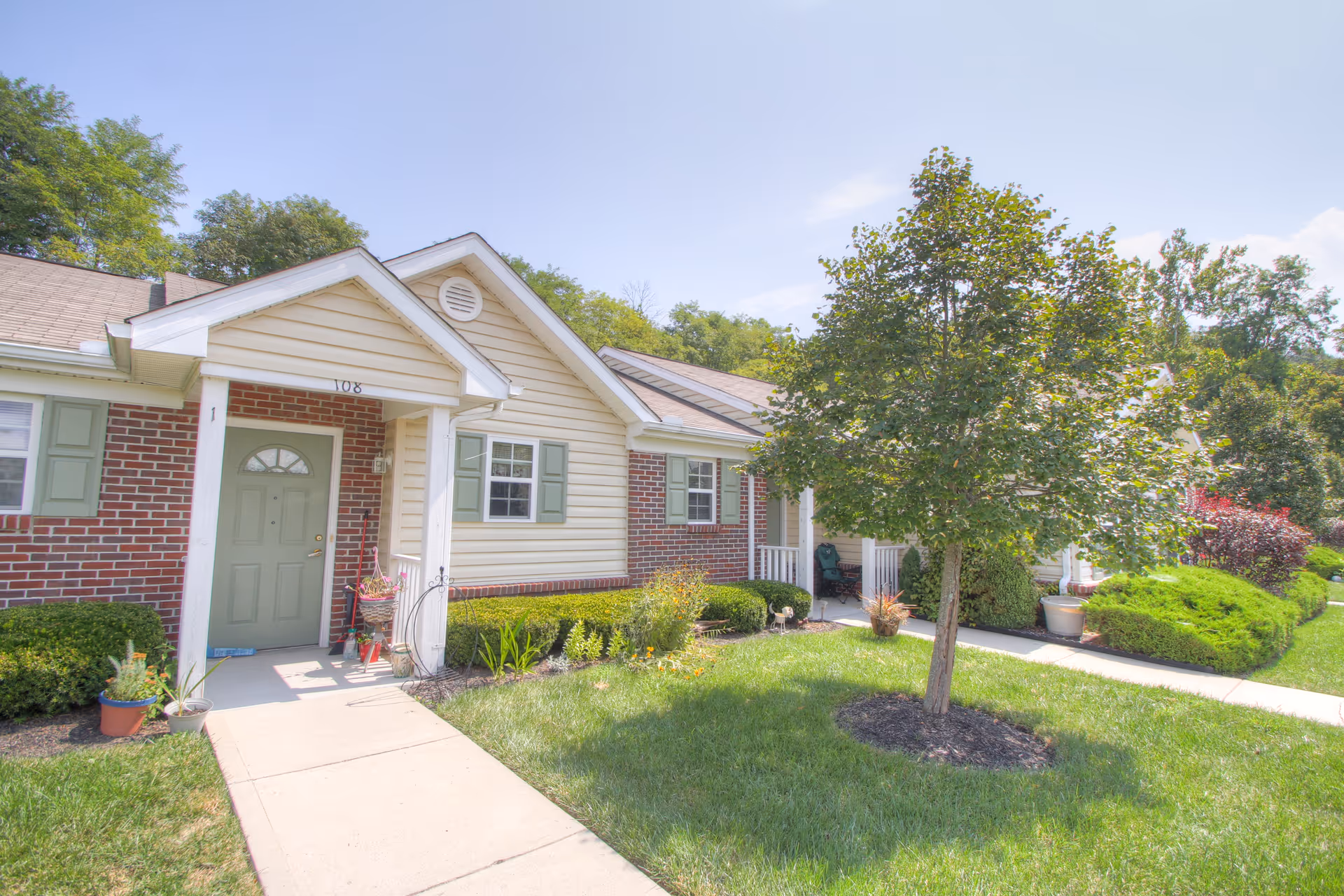 Exterior view of a single-story residential building with a green door, brick and beige siding, green shutters, and a small covered porch. There is a concrete walkway leading to the door, surrounded by green grass, bushes, potted plants, and a tree in the front yard under a clear blue sky.
