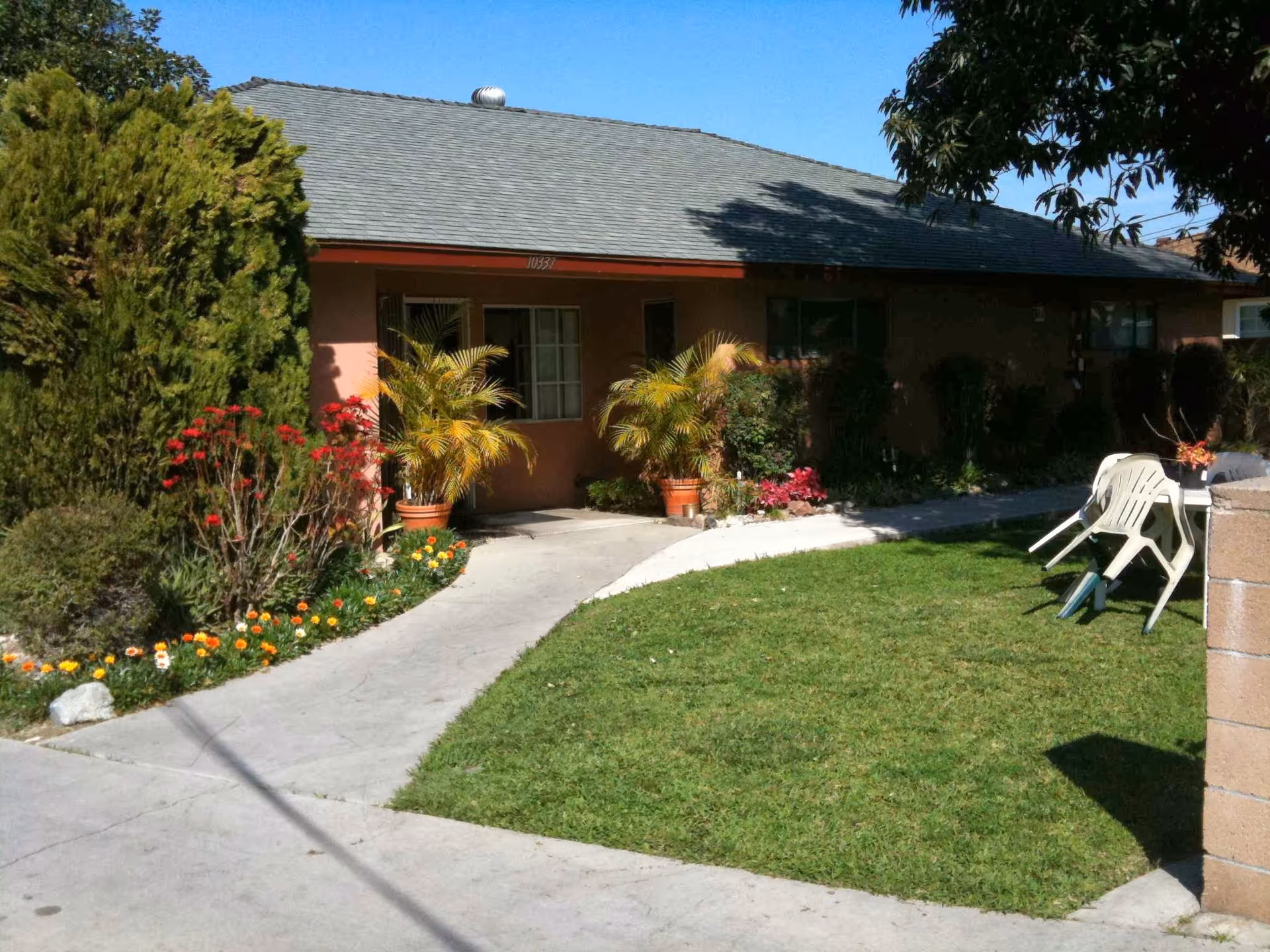Single-story residential building front with a paved walkway, lawn, potted plants and plastic chairs.