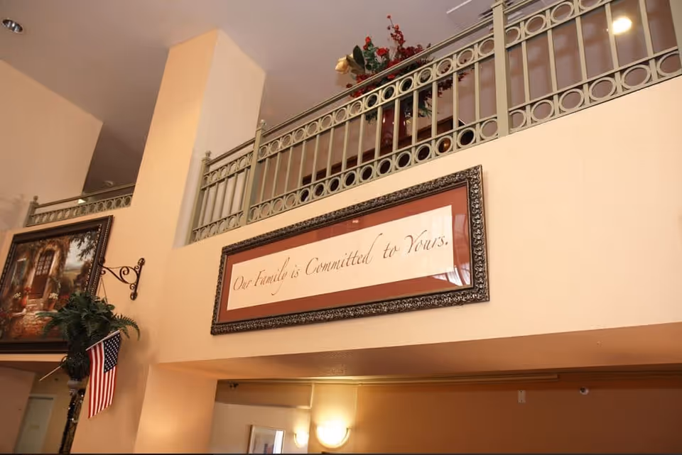 Interior lobby area with a decorative upper railing, a framed sign reading 'Our Family is Committed to Yours,' a painting, plant and a small American flag.