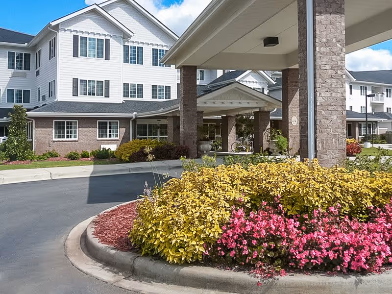 Exterior view of Shads Landing Gracious Retirement Living facility showing a covered entrance with brick pillars, a driveway, and landscaped flower beds with yellow and pink flowers in the foreground. The building has white siding and multiple windows under a partly cloudy sky.