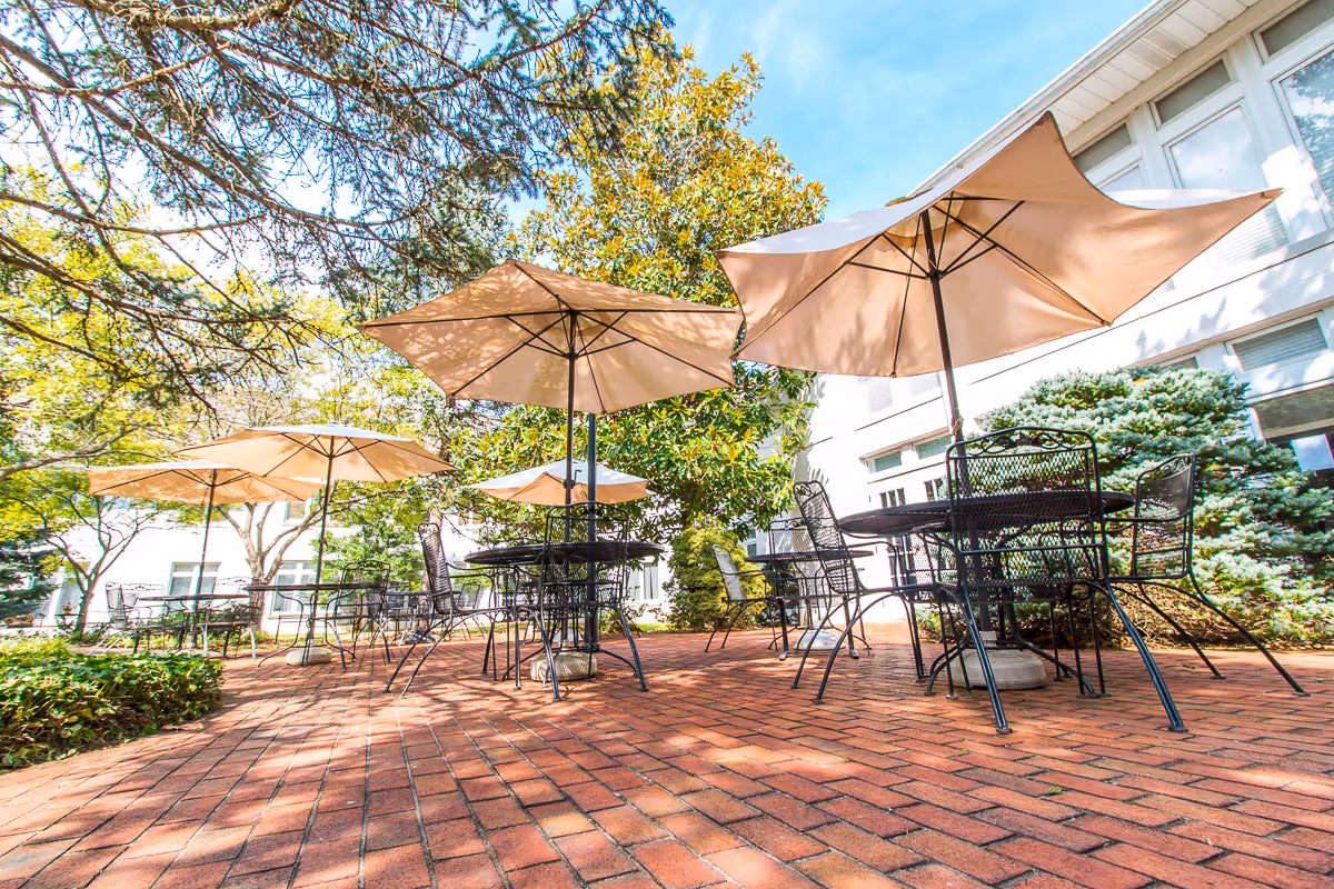 Outdoor patio area with several black metal tables and chairs, each shaded by large beige umbrellas. The patio is paved with red bricks and surrounded by green trees and shrubs, with a white building visible in the background under a clear blue sky.