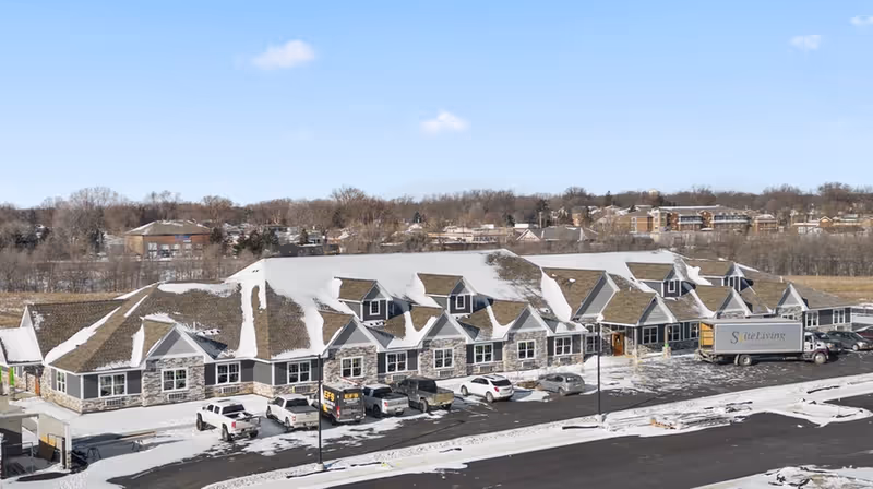 Snow-covered single-story senior living building with dormer windows, a row of parked cars and a delivery truck in front.