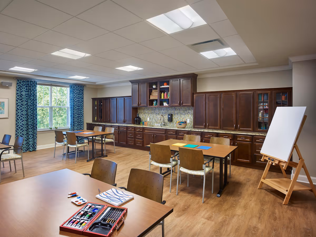 A well-lit activity room with wooden floors and multiple tables and chairs arranged for group activities. One table in the foreground has art supplies including paint tubes, brushes, and a palette. The back wall features dark wooden cabinets with a countertop and a sink. There is a large window with blue patterned curtains letting in natural light, and an easel with a blank canvas stands to the right.