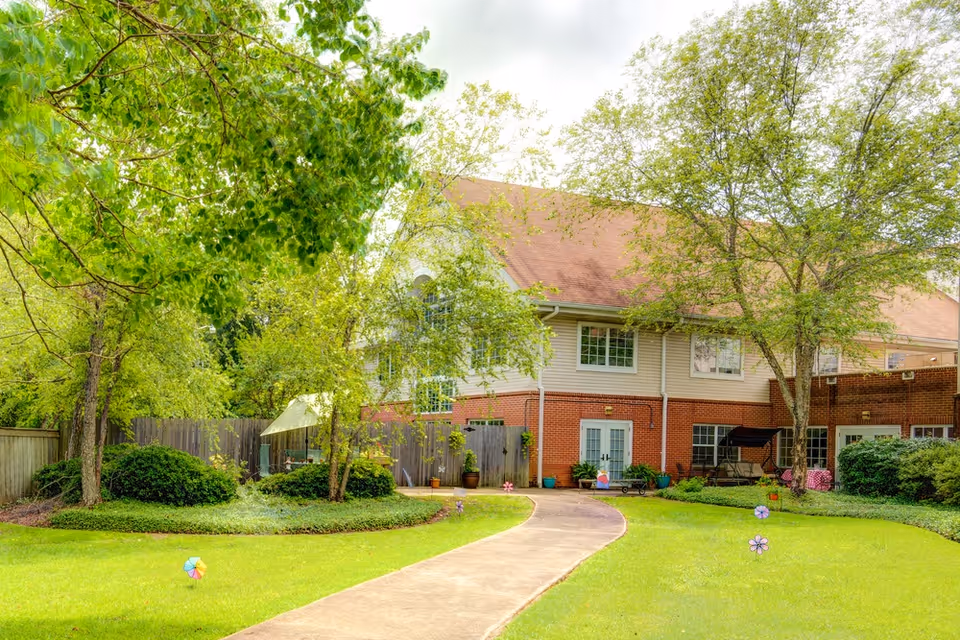 A peaceful outdoor garden area at Ridgeland Place with a curved concrete pathway, green grass, several trees, bushes, and a two-story building with a red roof and brick lower walls in the background. There are colorful pinwheels placed in the grass and a swing bench near the building.