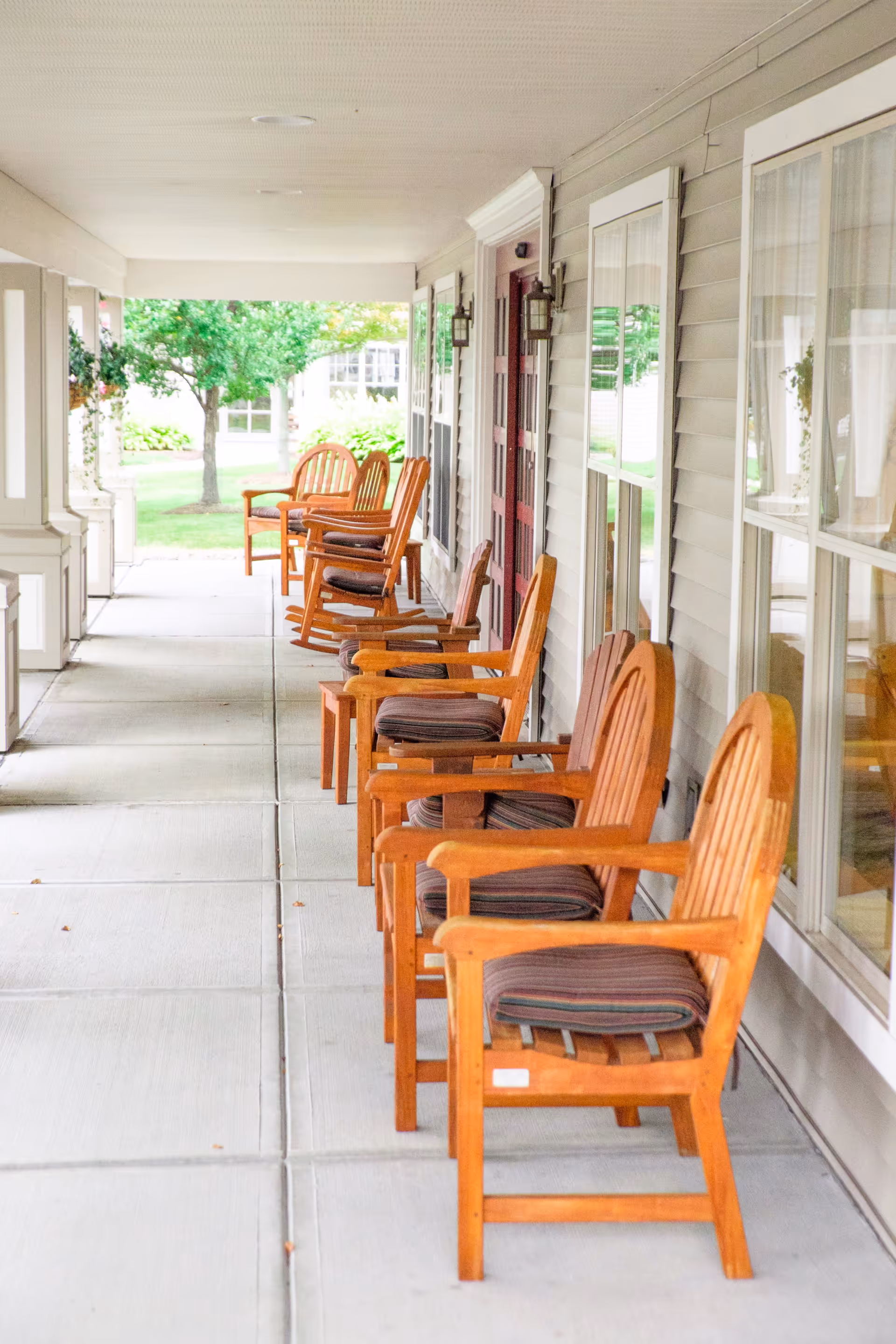 A covered outdoor porch area with a row of wooden chairs with cushions lined up along the wall of a building. There are windows and doors along the wall, and greenery including trees and bushes is visible at the end of the porch.