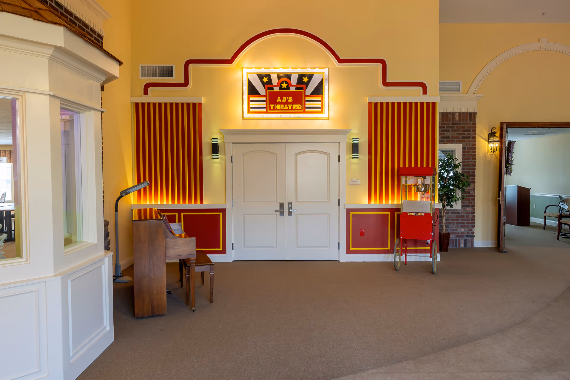 Interior view of a senior living facility hallway featuring double white doors labeled 'AJ's Theater' with illuminated marquee above. The walls around the doors are decorated with red and yellow vertical stripes and trim. To the left of the doors is a wooden piano with a bench and a floor lamp. To the right is a vintage-style red popcorn machine on wheels. The hallway has beige carpeting and light yellow walls with some brick accents and additional rooms visible in the background.