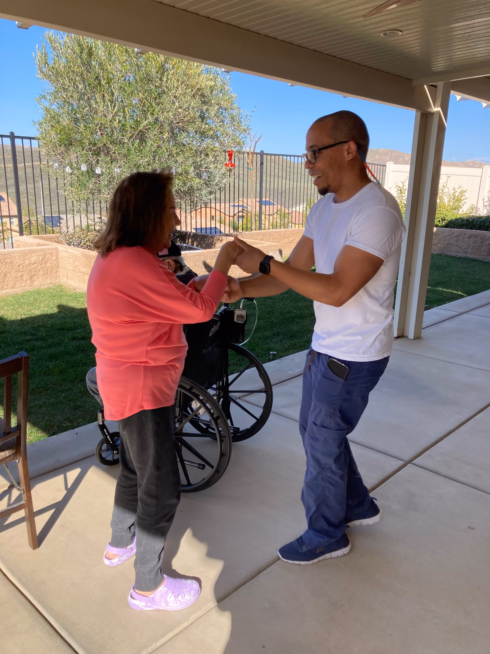 A woman in a pink top and gray pants is standing and holding hands with a man in a white t-shirt and blue pants under a covered outdoor patio. A wheelchair and a chair are nearby, and there is a grassy area with a tree and a fence in the background.