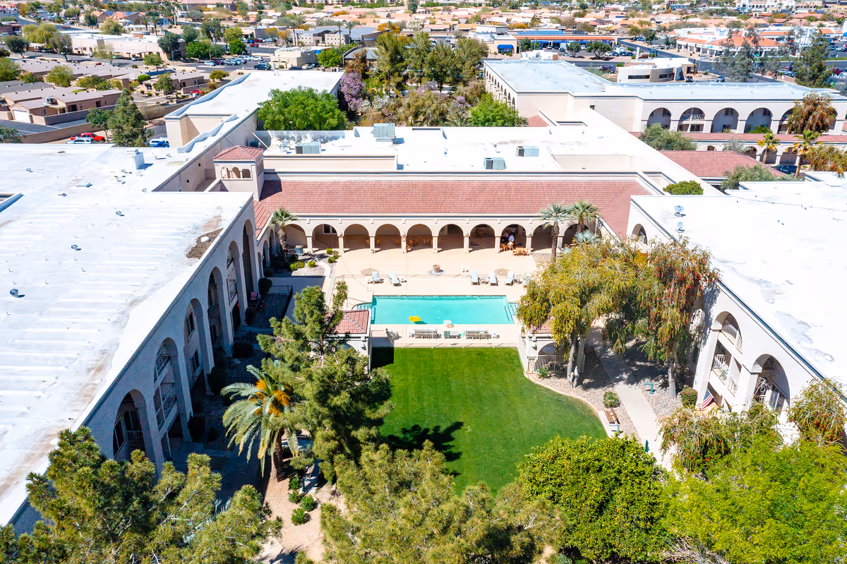 Aerial view of Solstice Senior Living at Sun City West showing a large courtyard with a swimming pool, surrounded by a two-story building with arches and a red-tiled roof. The courtyard has green grass, trees, and lounge chairs around the pool. The surrounding neighborhood is visible in the background.