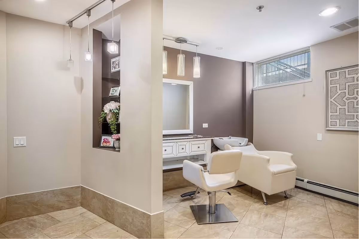 A modern salon area with a white styling chair and a white shampoo chair in front of a large mirror and countertop with drawers. The room has beige walls, a small window with blinds, tiled floor, and pendant lights hanging from the ceiling. There is a decorative wall niche with flowers and framed photos.