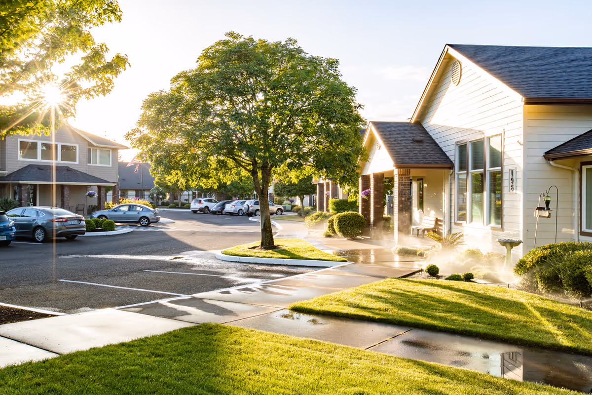 Sunlit exterior of a senior living community showing lawns, a tree-lined parking area, and single-story residences.