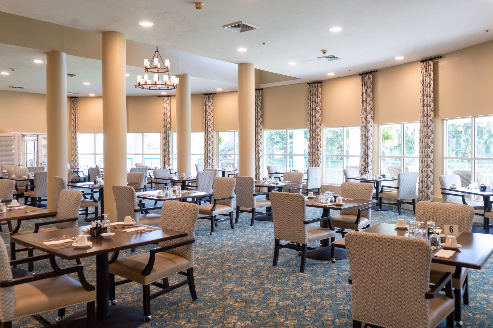 A spacious dining room with multiple tables and cushioned chairs arranged neatly. Each table is set with cups, glasses, and utensils. Large windows with patterned curtains allow natural light to fill the room, and there are several beige columns and a chandelier hanging from the ceiling.
