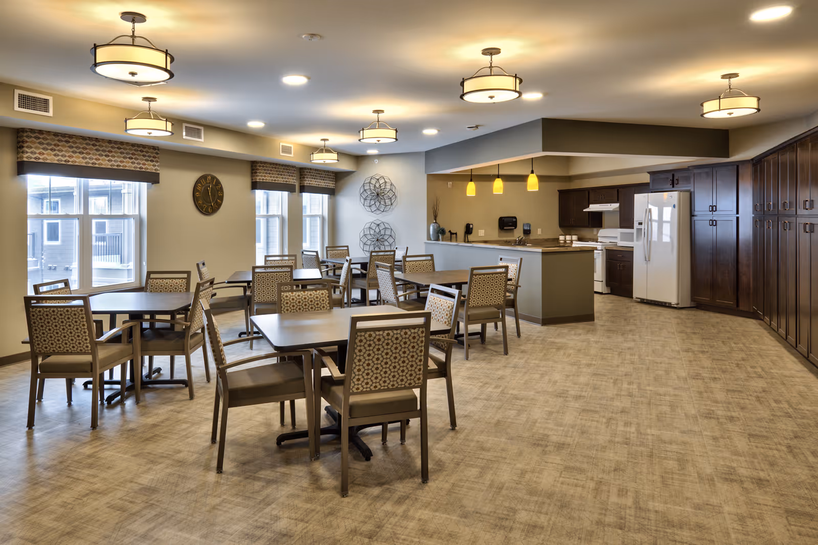 A spacious dining area in a senior living facility with multiple tables and chairs arranged neatly. The room features large windows with patterned valances, a clock on the wall, decorative metal wall art, and a kitchen area with dark wood cabinets, white appliances, and pendant lighting.