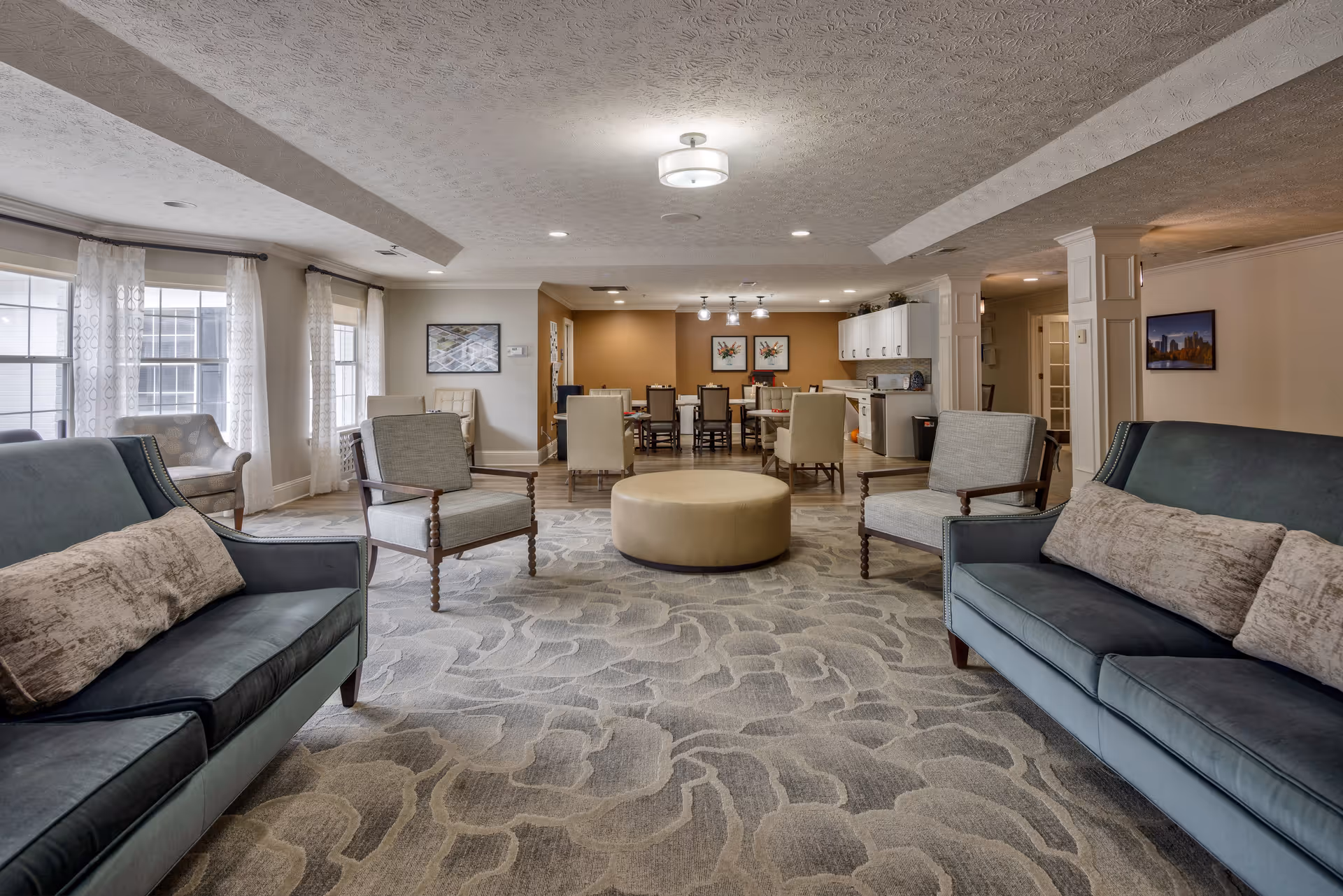 A spacious and well-lit common area in a senior living facility featuring two blue sofas with beige pillows, four wooden armchairs with cushions, a round beige ottoman in the center, and a dining area with tables and chairs in the background. The room has large windows with sheer curtains, patterned carpet, and ceiling lights.