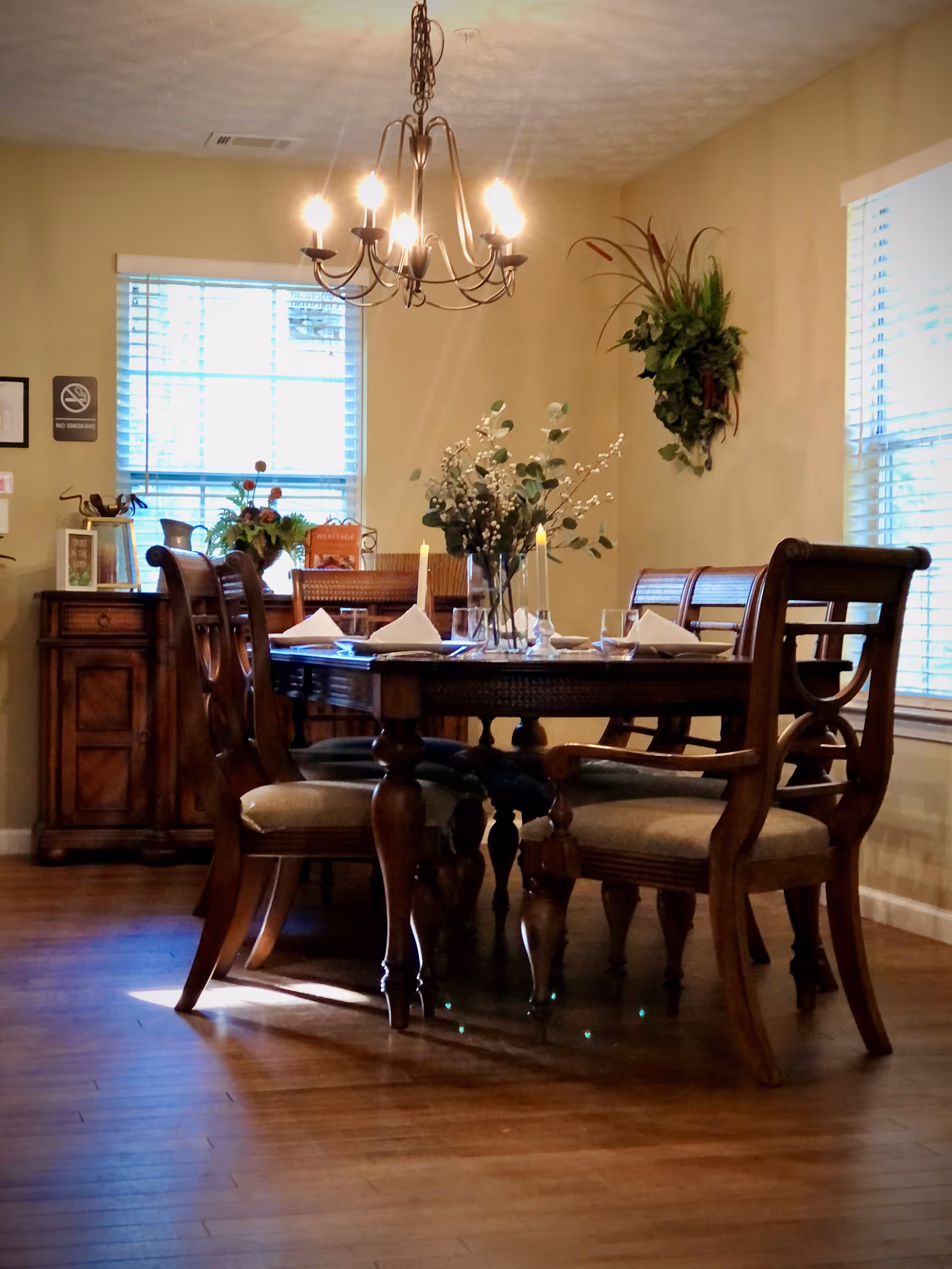 A warmly lit dining room with a wooden dining table set for four, including folded napkins, glasses, and candles. The room features wooden chairs with cushioned seats, a chandelier overhead, two windows with blinds, a sideboard with decorative items, and a wall-mounted plant arrangement.