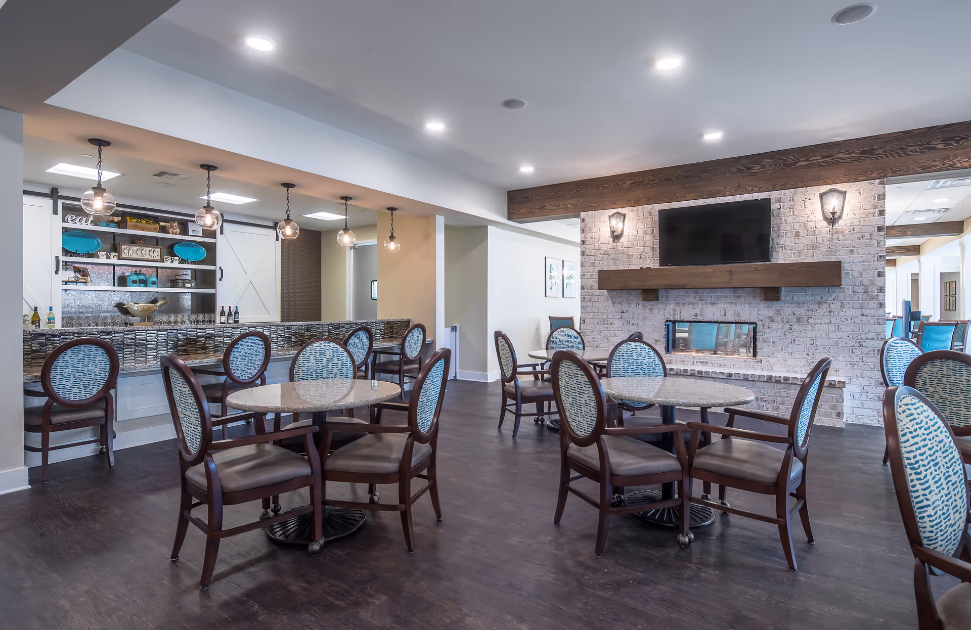 A bright and modern dining area with round tables and wooden chairs featuring blue patterned cushions. The room has dark wood flooring, a white brick fireplace with a mounted flat-screen TV above it, and a bar area with pendant lights and shelves displaying decorative items.