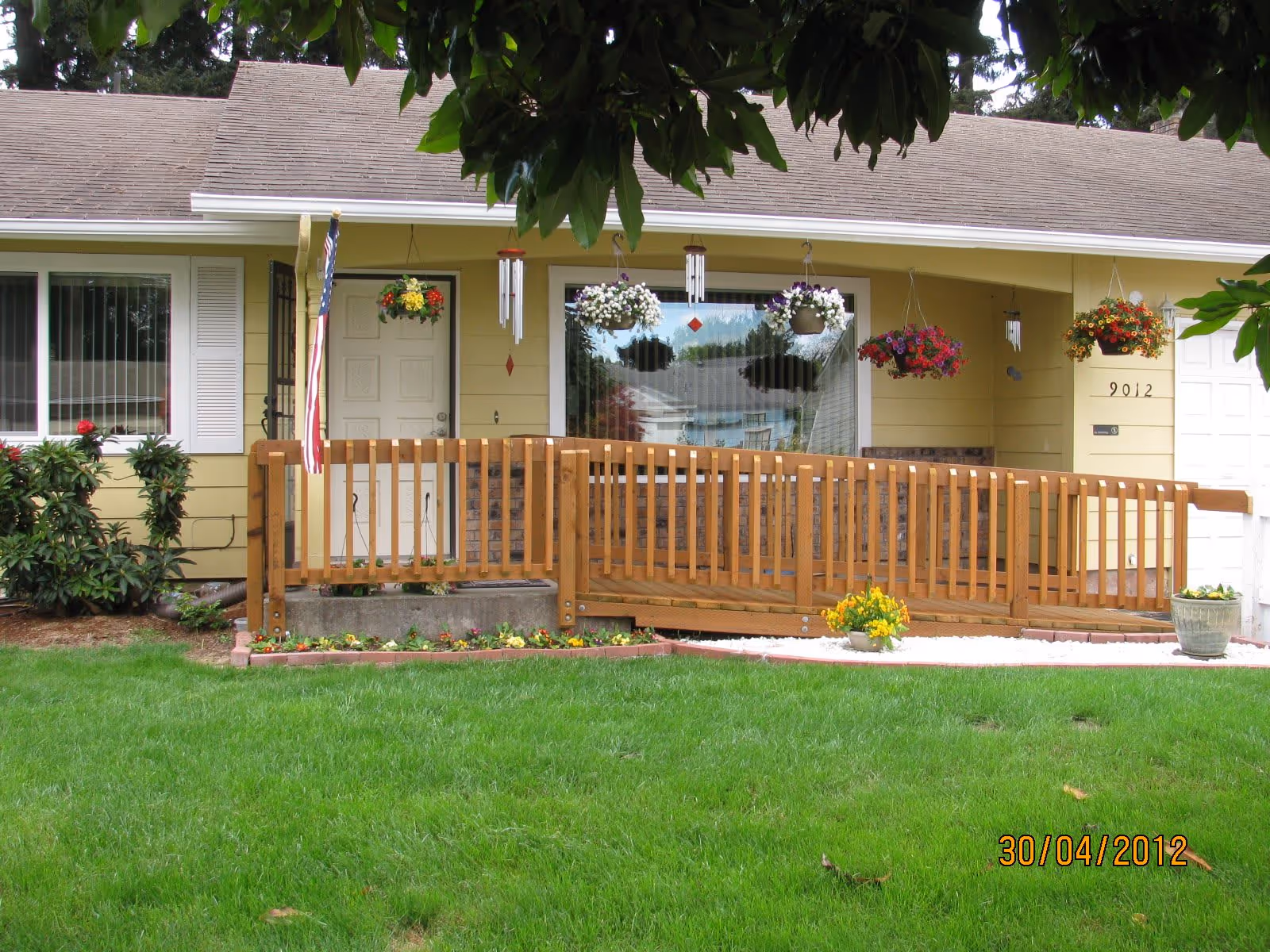 Front exterior view of a single-story yellow house with a brown shingled roof, a wooden ramp leading to the front door, hanging flower baskets, an American flag, and a well-maintained green lawn.