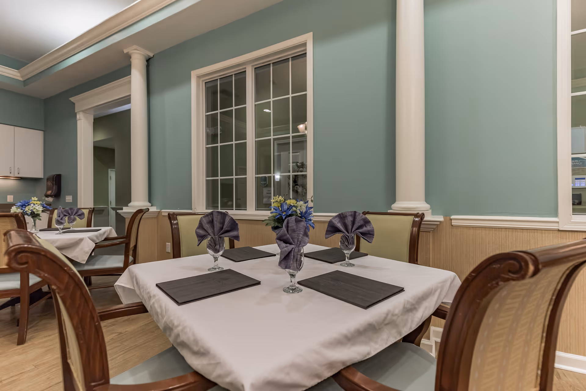 Dining area with tables covered in white tablecloths, each set with folded purple napkins in glasses and small flower arrangements. The room has light green walls, white trim, and wooden chairs with cushioned seats. There are large windows and decorative columns along the walls.