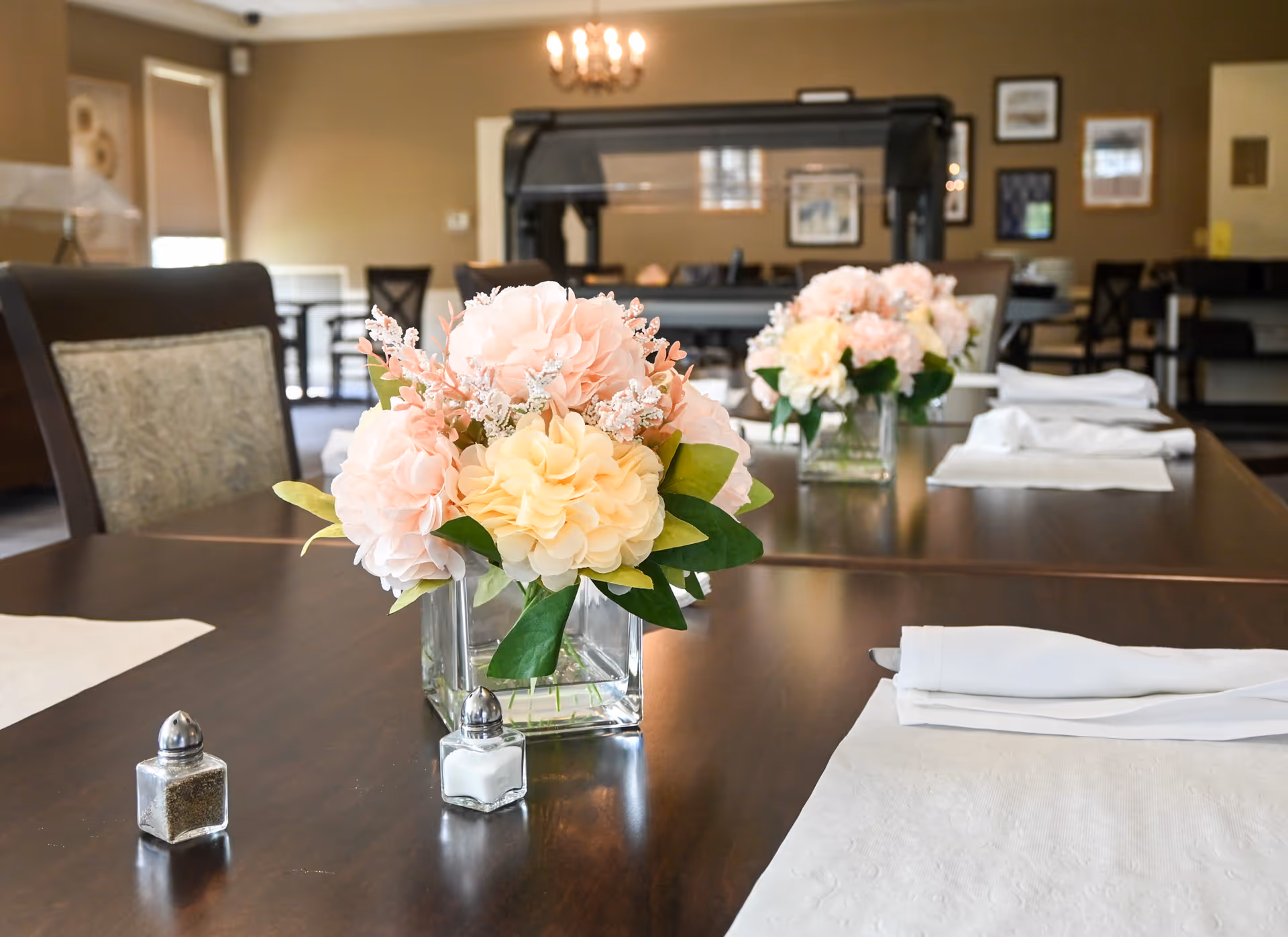 A dining table set with white napkins and placemats, decorated with a centerpiece of pink and yellow flowers in a glass vase. In the background, there are chairs and framed pictures on the wall in a softly lit room.
