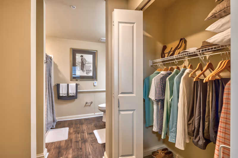 A walk-in closet with clothes on hangers opening into a bathroom with a toilet, shower curtain, and framed picture.