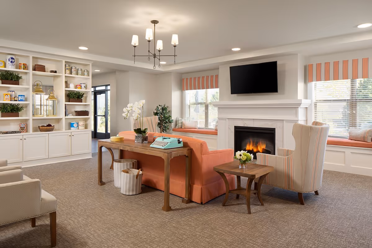 A cozy living room area with a coral-colored sofa, a striped armchair, a wooden coffee table with a small flower vase, and a fireplace with a mounted flat-screen TV above it. The room features built-in white shelves with decorative items and plants, large windows with striped valances, and a wooden console table behind the sofa with a vintage turquoise typewriter and a white orchid plant.