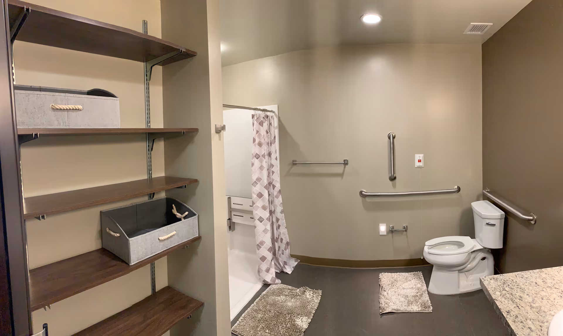 Accessible bathroom with a toilet, grab bars on the walls, a shower with a patterned curtain, and wooden shelves with storage bins. The floor has two small rugs and the walls are painted in neutral tones.