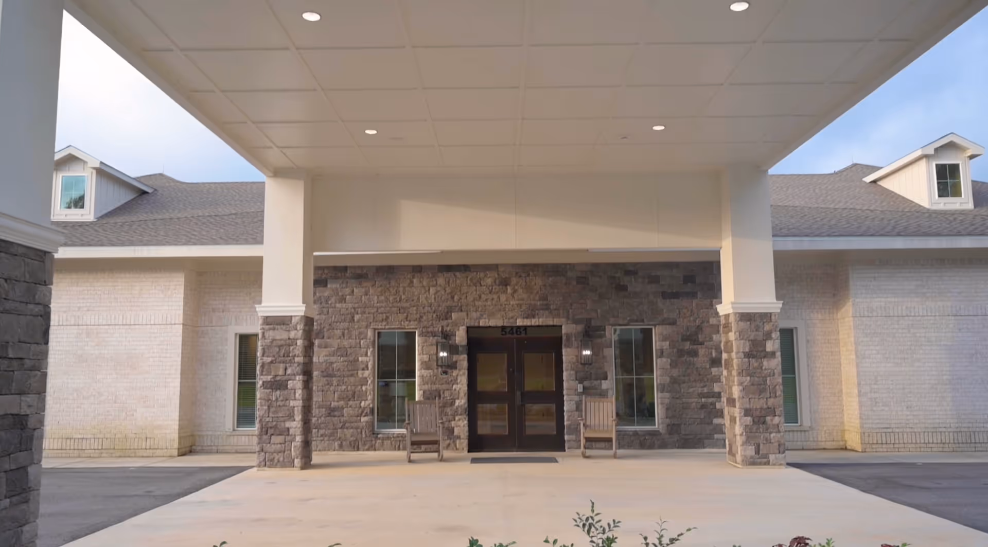 Entrance of a senior living facility with a covered drop-off area supported by stone pillars, two wooden chairs on either side of double glass doors, and a building facade made of light-colored brick and stone.
