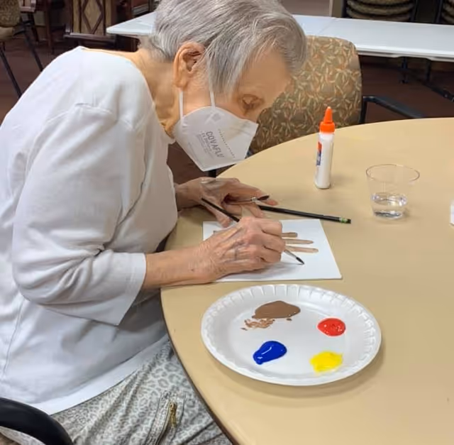 An elderly woman wearing a white face mask and white long-sleeve shirt is sitting at a round table, painting on a piece of paper. On the table are a paper plate with blobs of blue, brown, red, and yellow paint, a bottle of glue, a pencil, and a small clear cup of water. The setting appears to be an indoor common area with chairs and tables.